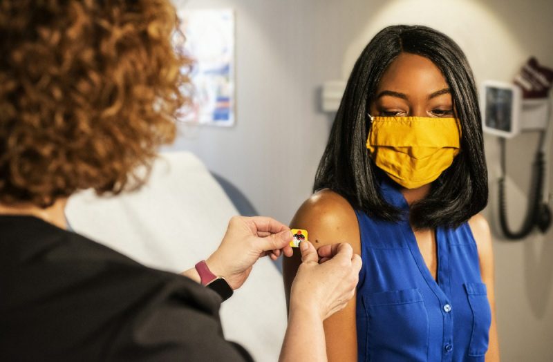 Young Black woman wearing a yellow face mask receiving a band aid after a vaccine.