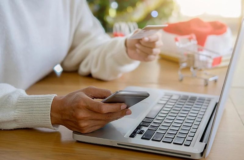 A person's hand holding a phone and credit card sitting in front of a laptop.