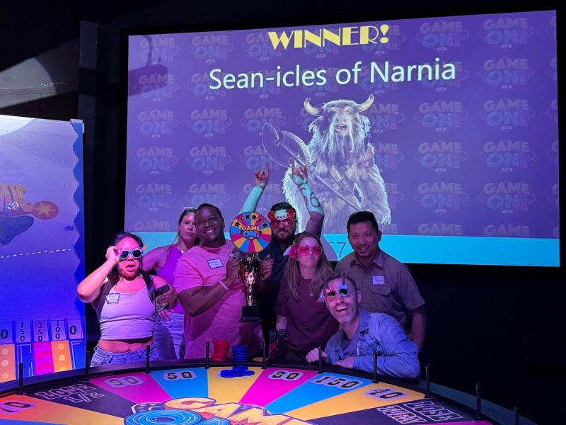 a group of smiling people stand behind a wheel of fortune type wheel